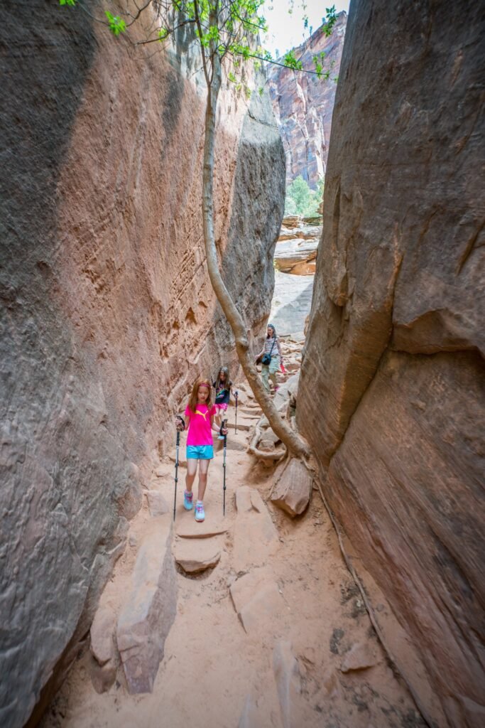 Hikers navigating a tight, narrow sandstone slot canyon with towering rock walls and a sandy path.