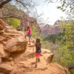 Canyon Overlook Trail Hikers with trekking poles navigating a rocky trail through a deep red rock canyon. Lush green trees and shrubs line the path, reflecting the area's natural beauty.