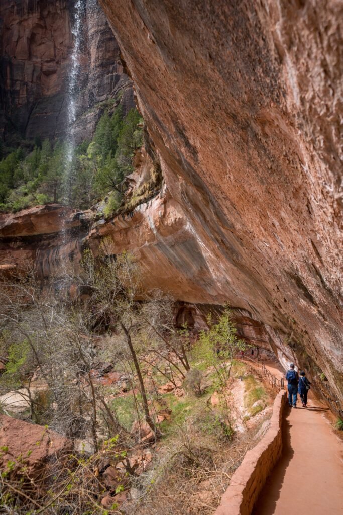 Hikers walking a dirt trail in red sandstone canyon with waterfall above