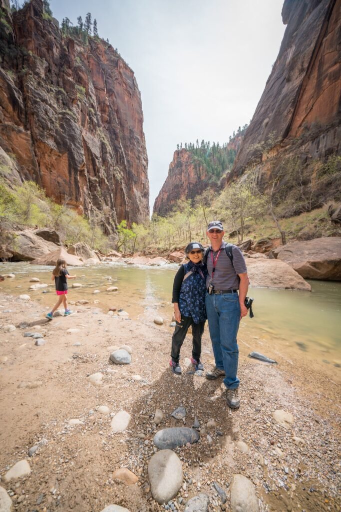 Two hikers pose beside a river along a rocky canyon trail. A third person is seen walking nearby as towering cliffs rise overhead.