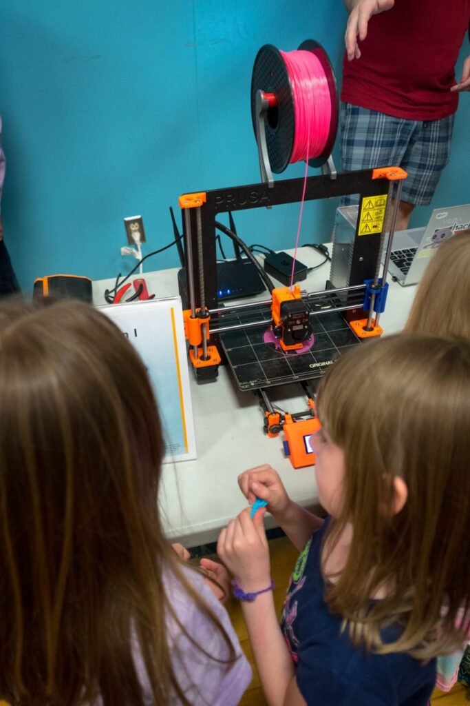 Children observe a 3D printer with pink filament at a white table against a teal wall.