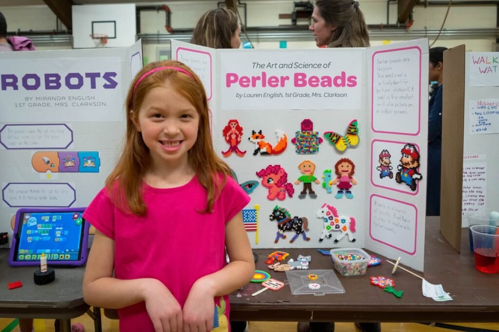 A student in a pink shirt presents a science fair board titled 'The Art and Science of Perler Beads', with colorful bead creations and a display table featuring a digital screen.