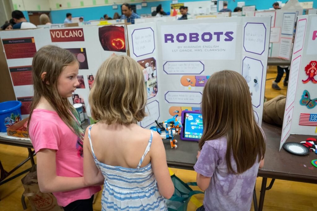 Children investigate a robots demonstration table at an outdoor science fair. Nearby displays feature topics on volcanoes and other scientific concepts.