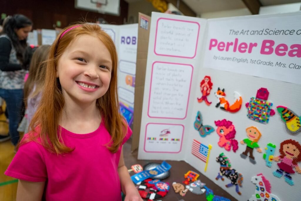 A smiling child in a pink shirt stands next to a science fair display board featuring colorful Perler bead creations. The tri-fold poster highlights the theme “The Art and Science of Perler Beads.”
