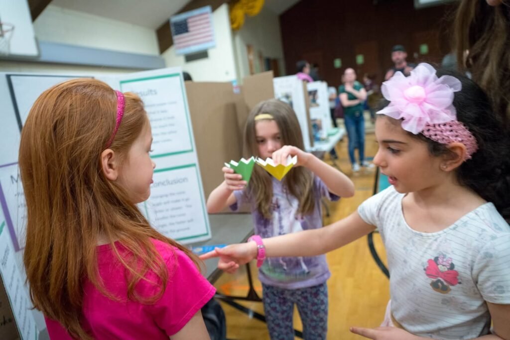 Two young girls interacting at an indoor event while another holds a colorful paper craft in the background.
