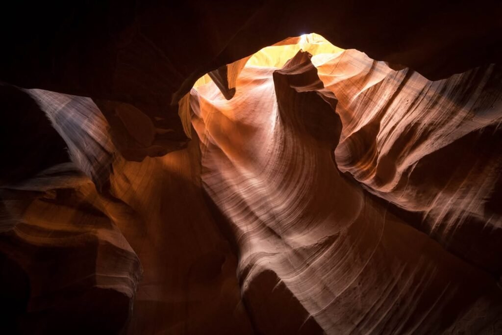 Sunlight illuminates dramatic formations inside a narrow slot canyon. Wavy sandstone walls display vibrant geological layers in warm tones.