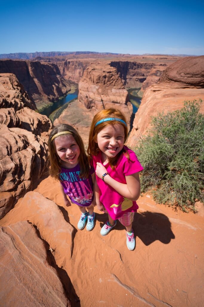 Two children standing on a red rock formation overlooking a vast canyon with a teal river under a clear blue sky.