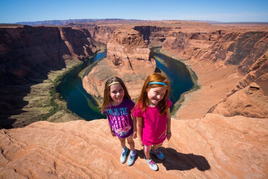 Two visitors stand on the edge of the Grand Canyon, capturing the stunning red rock formations and vibrant blue river. The panoramic view highlights the vast expanse of this natural wonder.