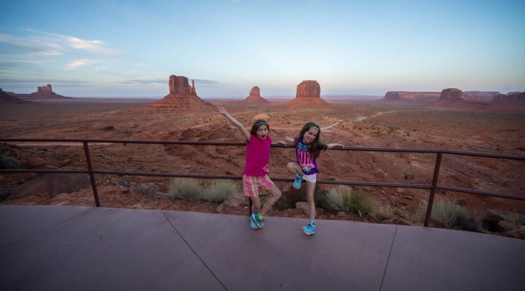 Two people stand on a scenic overlook with Monument Valley buttes in the background.