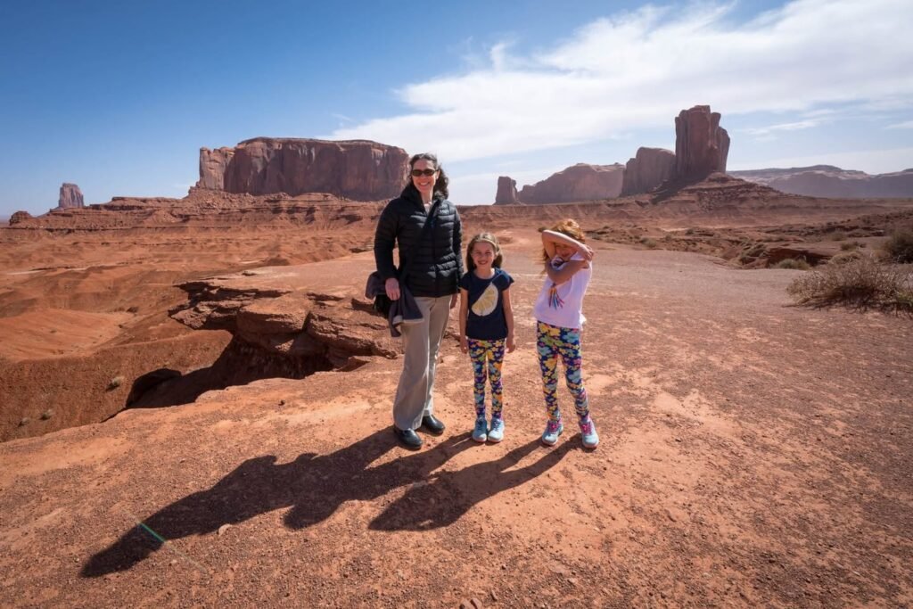 Three people standing on red rock terrain in a desert landscape with prominent rock formations in the background. The scene features a wide, arid setting under a blue sky with scattered clouds.
