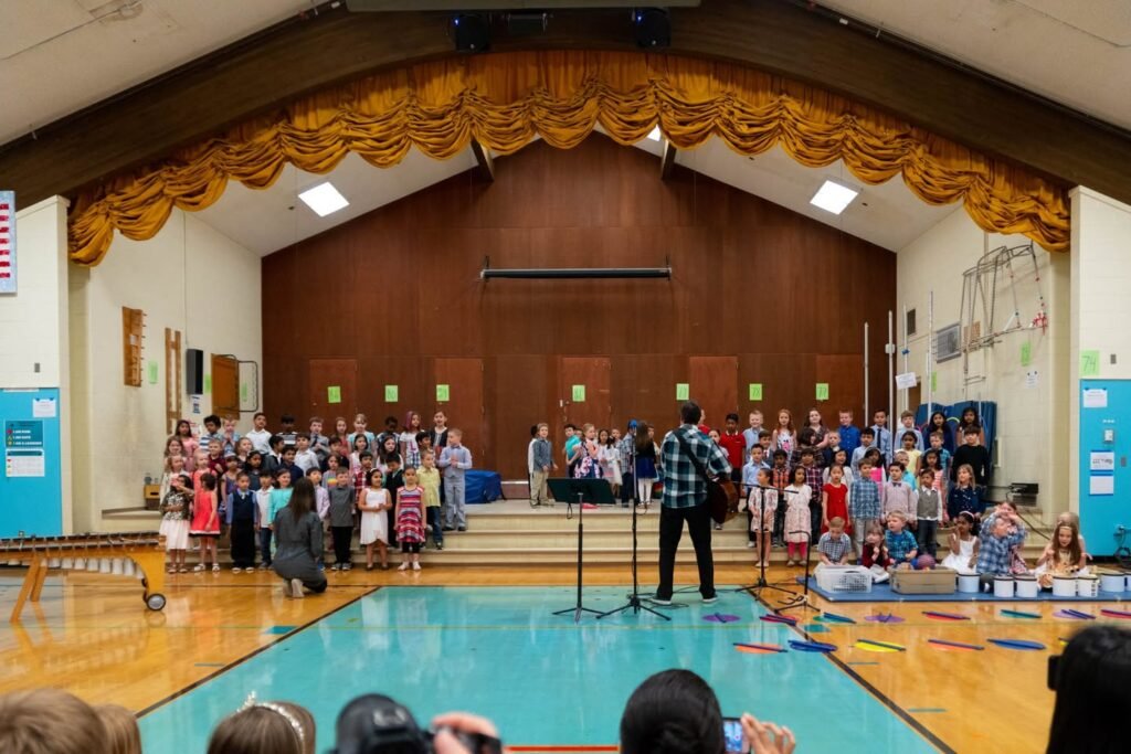 An indoor community event in a wooden gymnasium with a large audience and an empty pool.