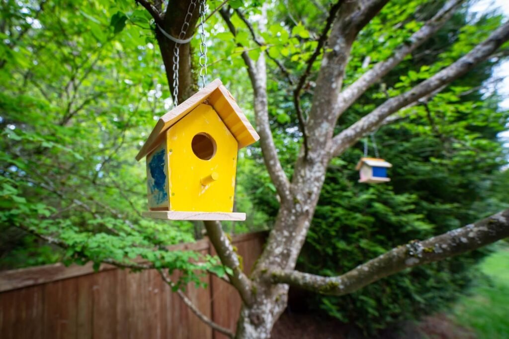 A yellow birdhouse with a wooden roof and perch hangs from a tree in a lush green garden.