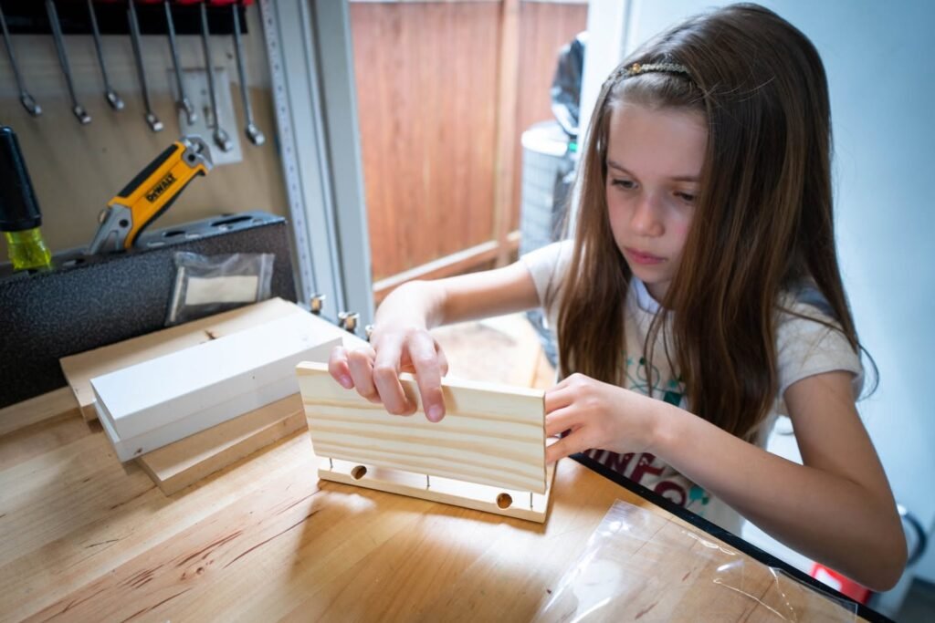 A person assembling a wooden object at a workbench with power tools in the background.