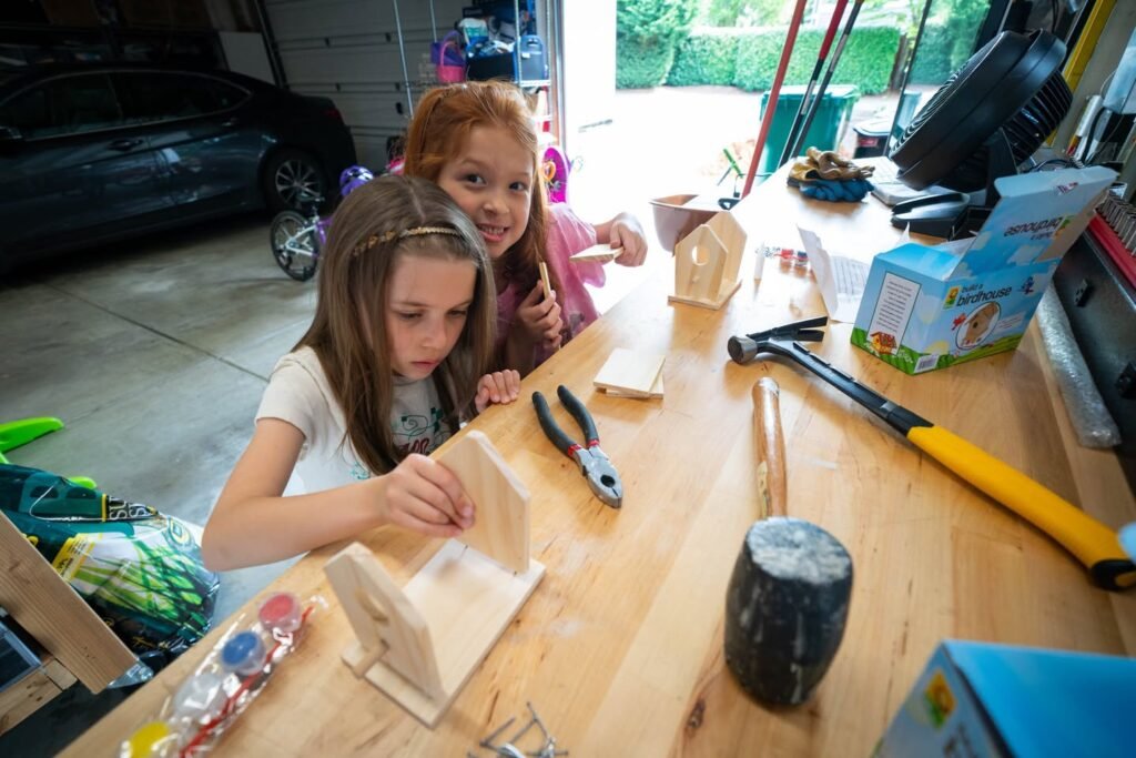 Children working on a woodworking project at a garage workbench, surrounded by tools and a car in the background.