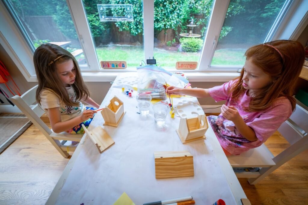 Two children engaged in a craft project at a table near a large window with a view of lush greenery outside.