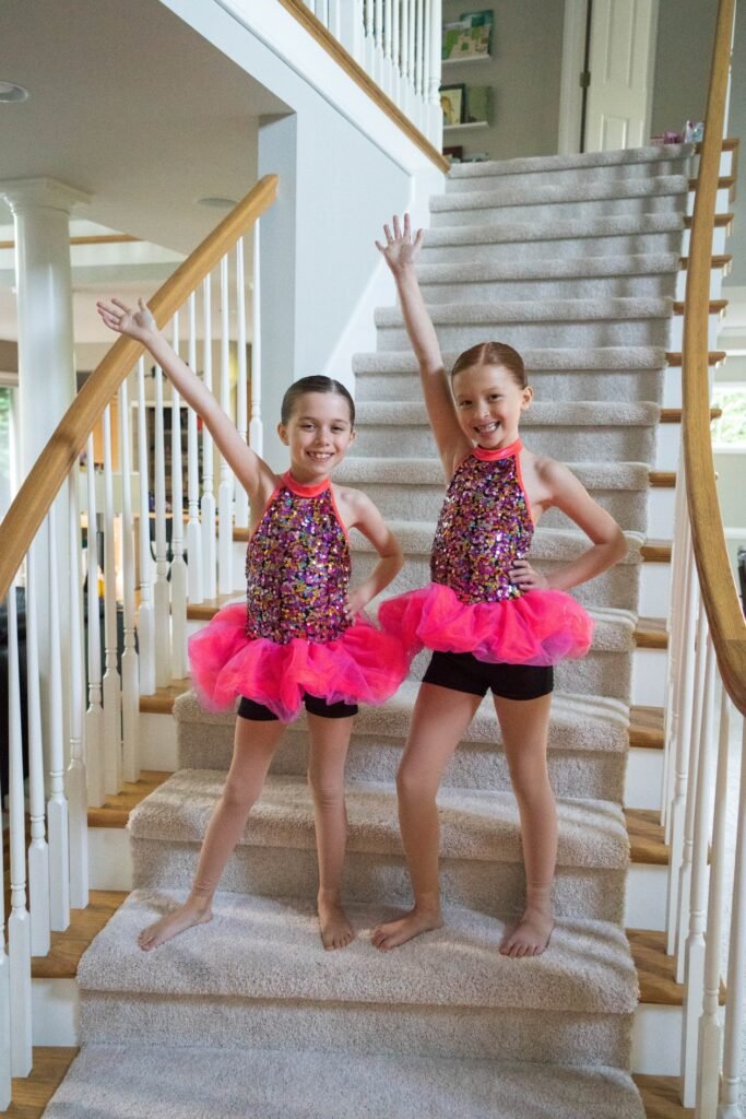 Children wearing colorful sparkly tops and pink tutus pose on a carpeted staircase with one arm raised.