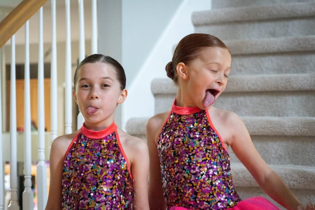 Two children in colorful sequined gymnastic leotards sit on stairs making silly faces.
