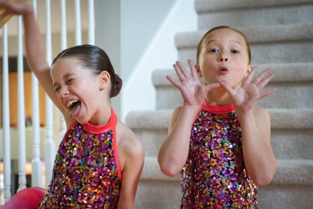 Two children in sparkly multi-color sequin dresses posing on a carpeted staircase with a white railing, one laughing with arm raised, the other making a face and waving hands.