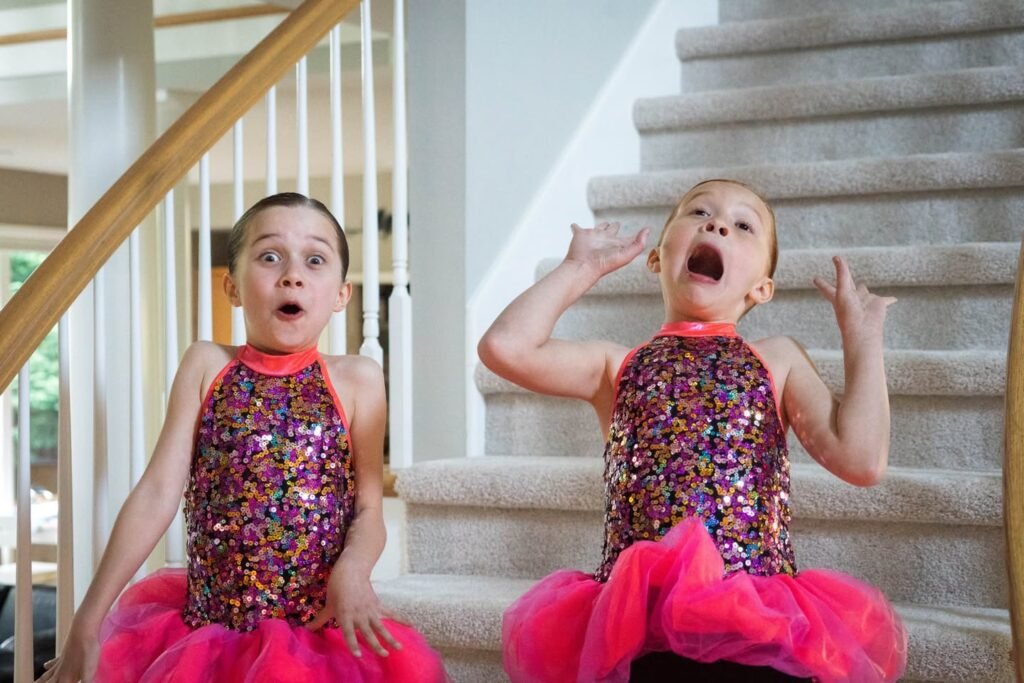 Young children wearing sequined dance tops and bright pink tutus standing on a carpeted staircase with surprised expressions.