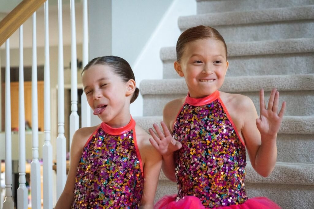 Two young dancers in matching sequined costumes waving from a home staircase.