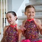 Two young dancers in matching sequined costumes waving from a home staircase.