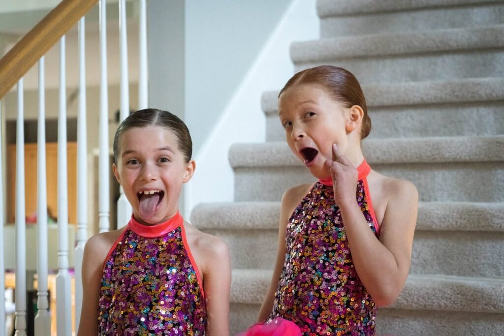 Children in purple sparkly leotards playfully pose on carpeted stairs. One extends tongue for a silly face while another shows surprise.