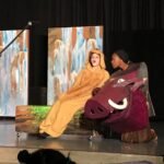 Jupiter A young performer in a yellow robe next to a person in a pig costume on a stage.