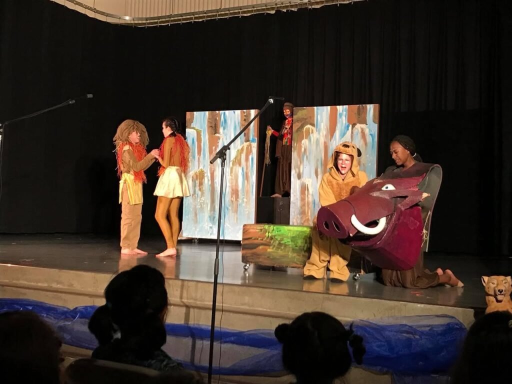 Performers in colorful costumes on stage, featuring a large animated bear prop and a scenic backdrop. A pupil audience is visible in the foreground watching the show.
