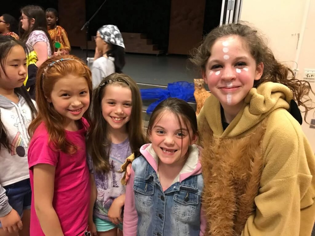 A group of young girls smiling after a performance, with one wearing face paint in the foreground.