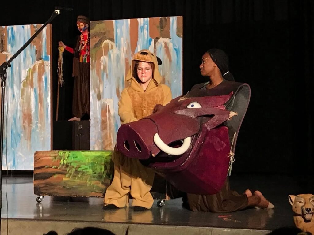 Performers in colorful costumes present a scene on a stage with painted backdrops and a large maroon prop.