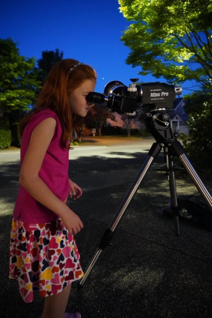 A person peers through a telescope mounted on a tripod against a dusk sky. Leaves are visible in the background.