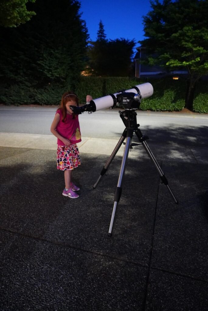 A telescope on a tripod set up in a driveway at night for stargazing, with a clear blue dusk sky.