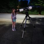A child peers through a telescope set up on a tripod during twilight hours.