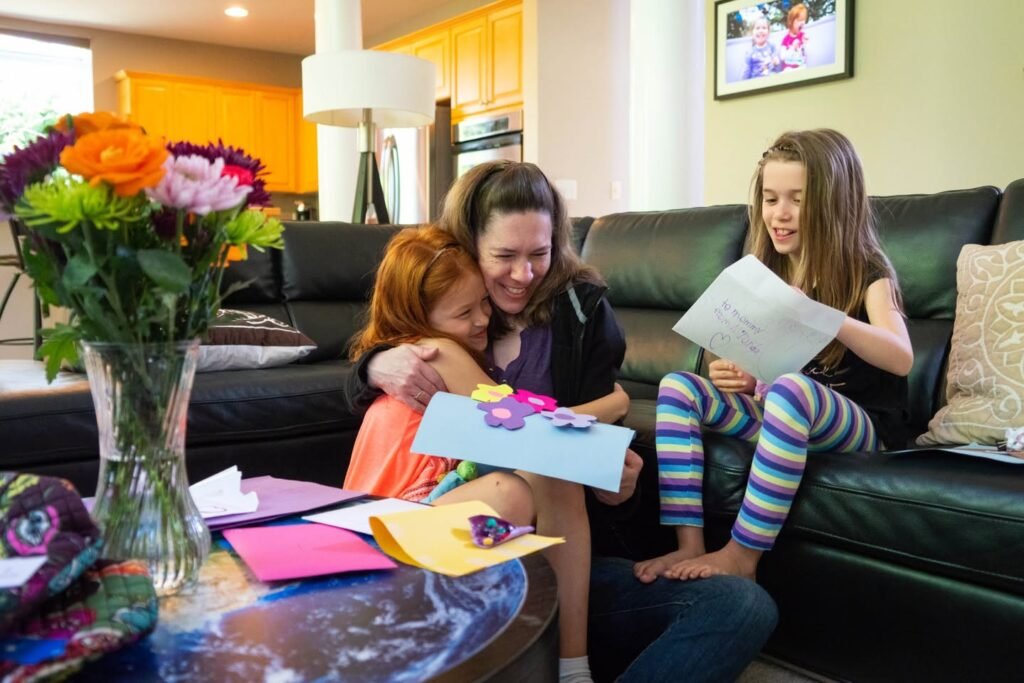 A woman embracing a child on a couch while a girl reads a card, with a vibrant flower arrangement in the foreground.
