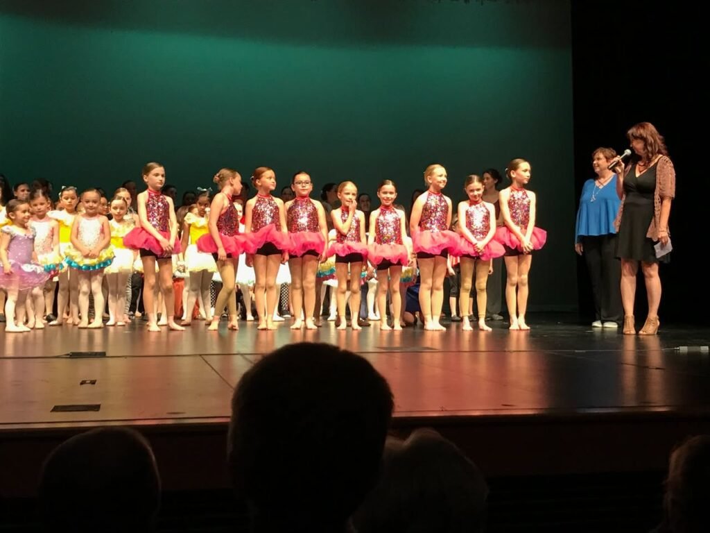A group of young dancers in colorful tutus and leotards on stage during a recital, with an audience perspective in the foreground.