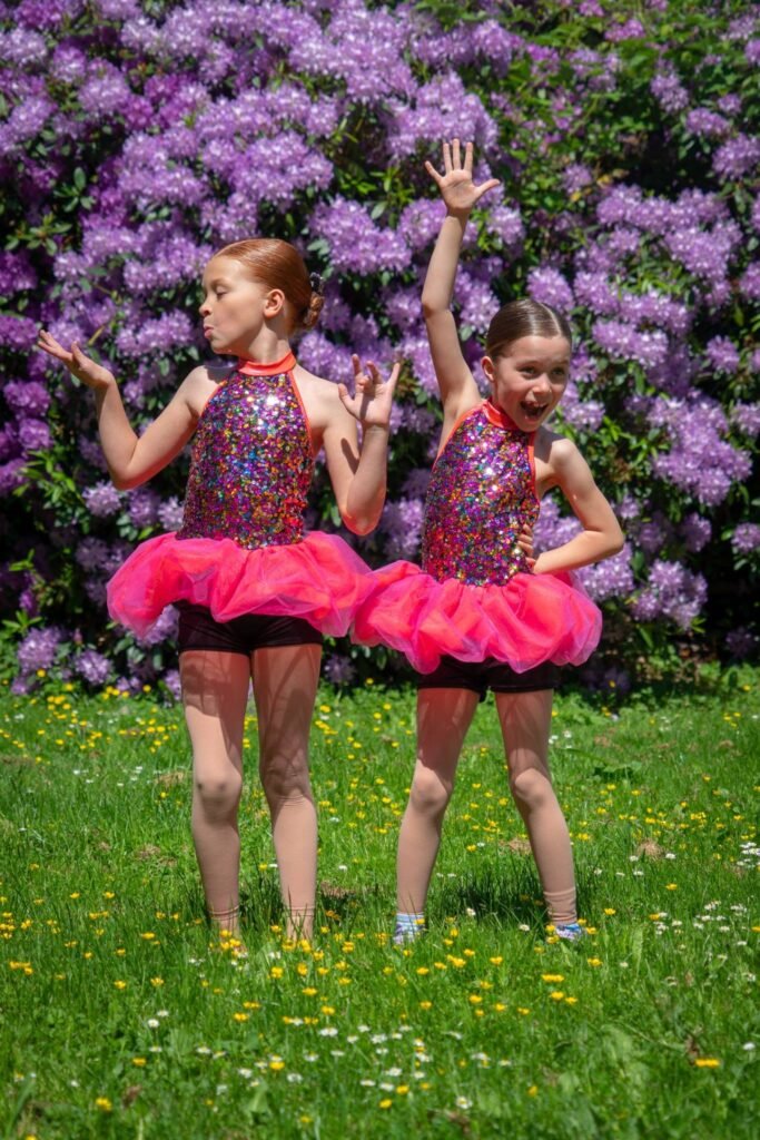 Two dancers in pink sparkly outfits perform in front of lush purple flowers on a sunny day in the Pacific Northwest.