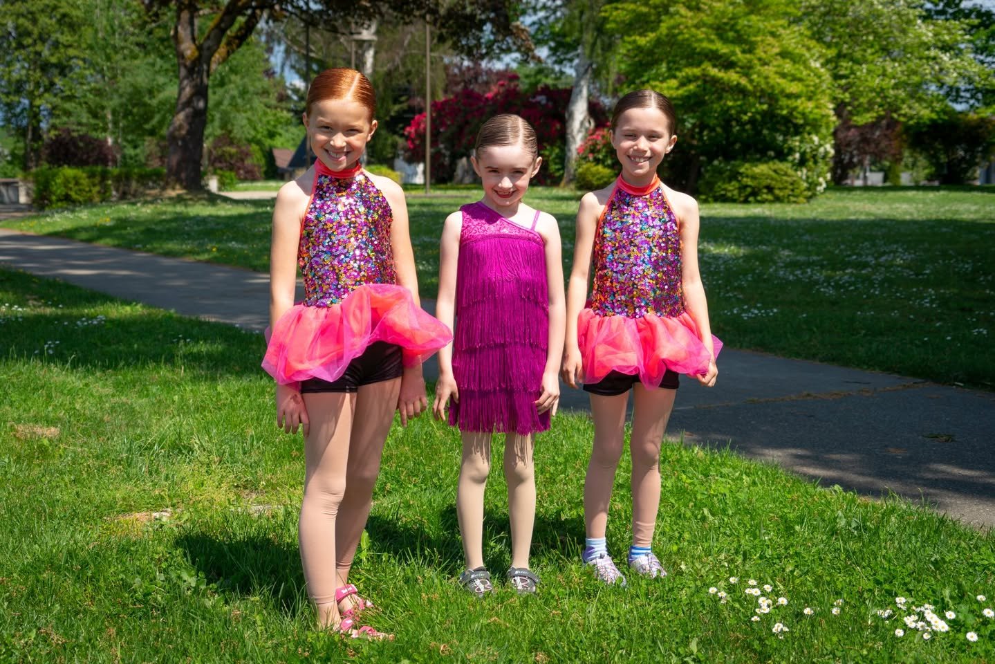 2018 Dance Recital Three people in colorful dresses stand on the grass in a park setting with trees in the background.