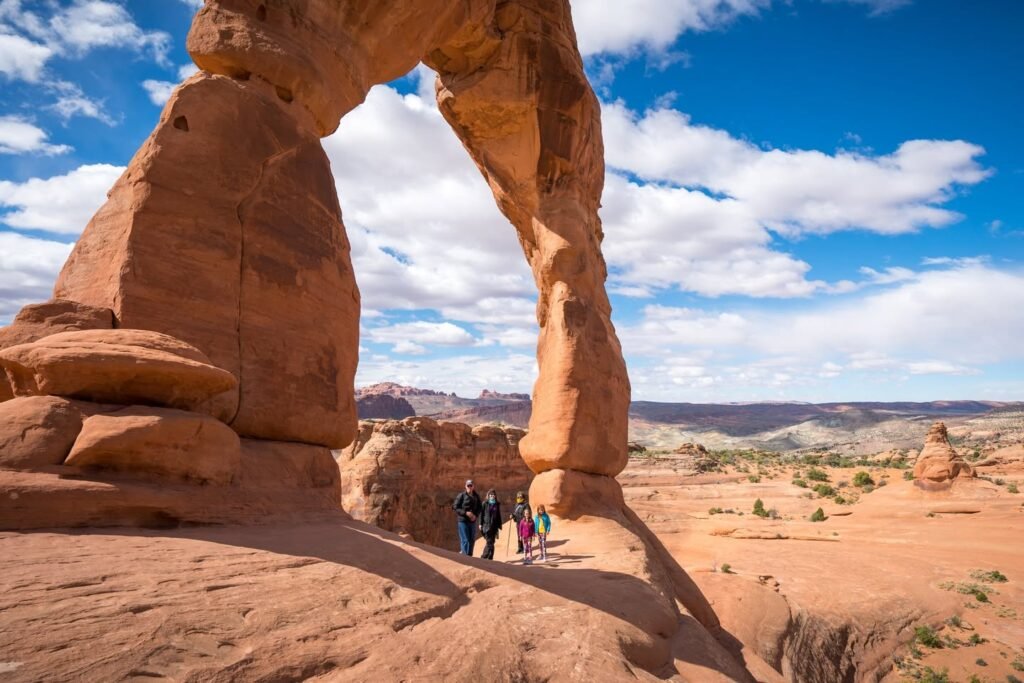 A group of hikers stands beneath a towering sandstone arch in a desert landscape.