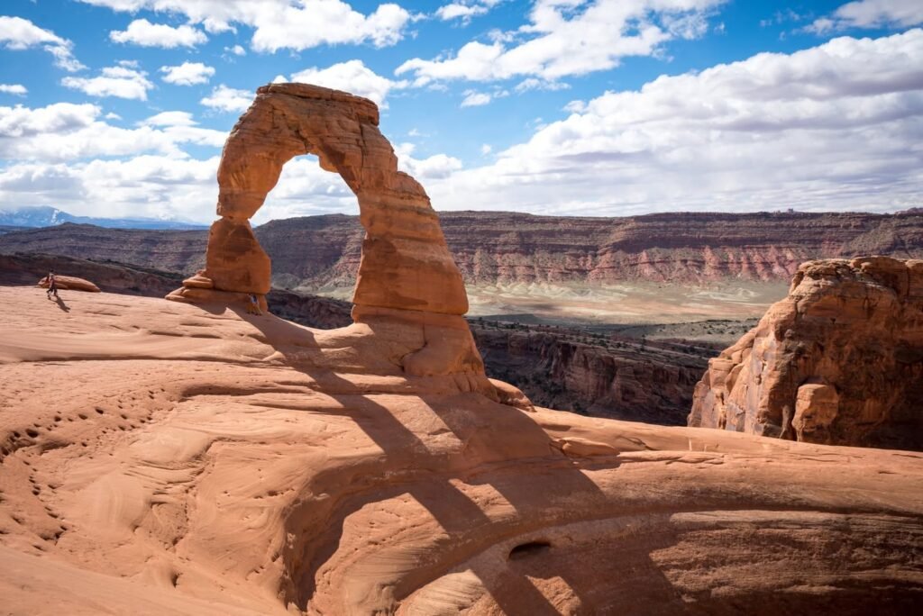 A stunning natural rock arch stands tall in a rugged desert landscape with a vibrant blue sky and scattered clouds.