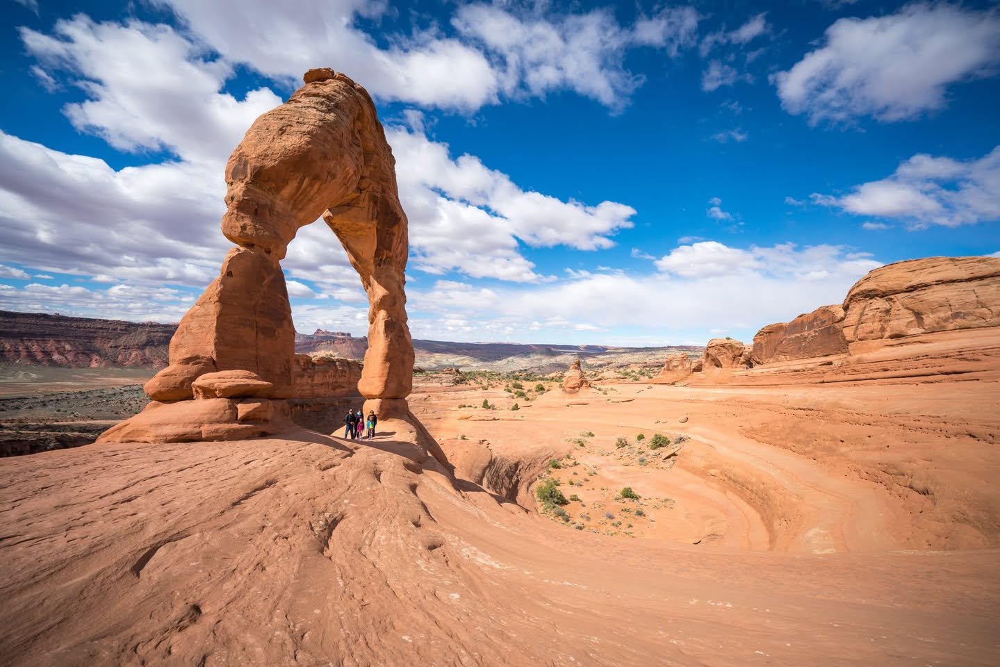 Spring Break – Delicate Arch A panoramic view of red rock desert formations beneath a vibrant blue sky. The natural arches and wind-carved sandstone dominate the scene, showcasing the raw beauty of the arid environment.