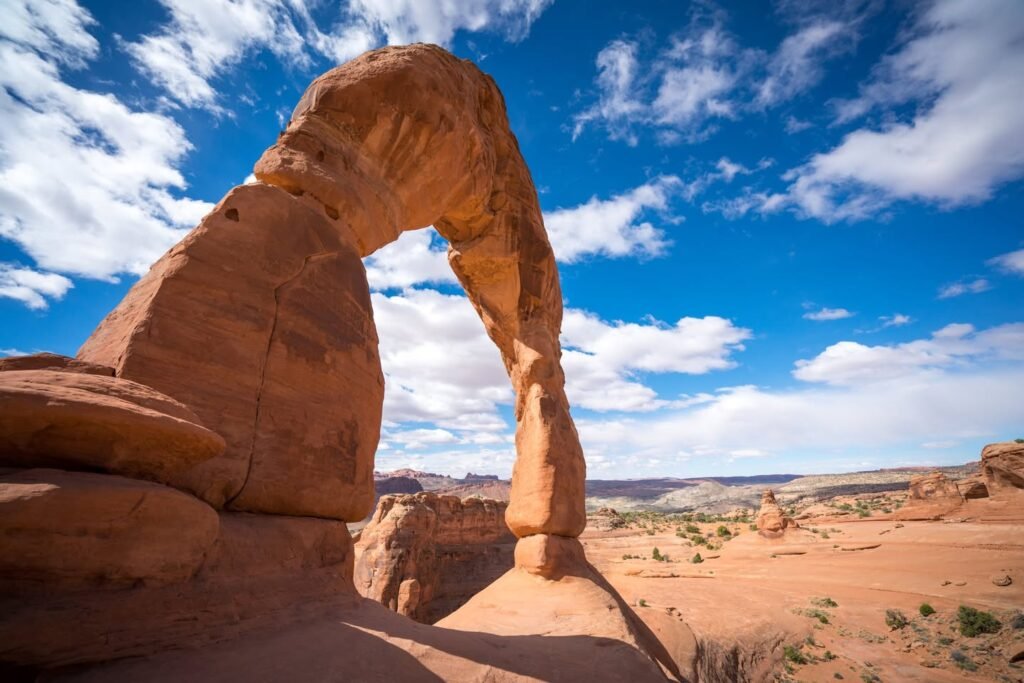 A natural rock arch stands tall against a vibrant blue sky with scattered clouds. The desert landscape below features rolling sandstones and sparse vegetation.