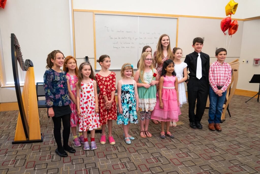 A group of children dressed in formal and semi-formal attire standing in a classroom next to a harp during a music recital or concert.