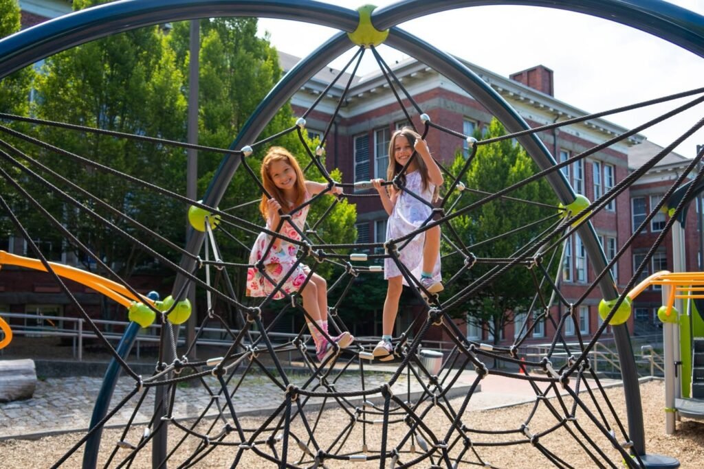 Children climbing on a netted playground structure with a brick building and trees in the background.