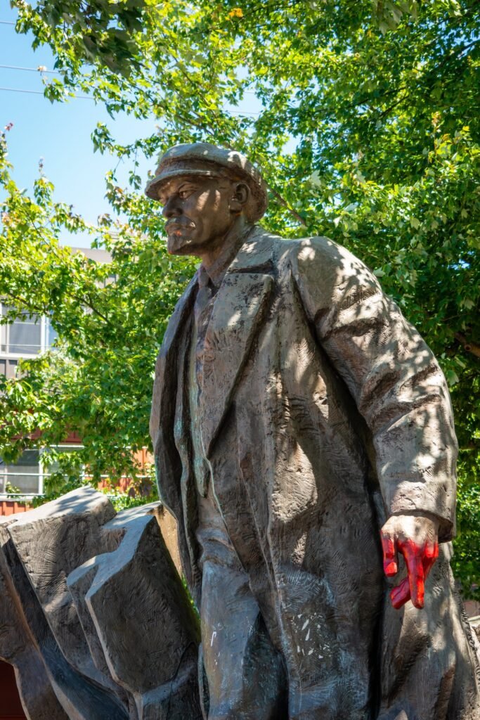 A detailed bronze statue of a man in a coat and cap, holding a red object, set against a backdrop of lush green trees and clear blue sky.