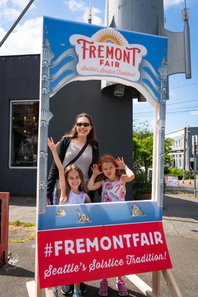 A family posing in a decorative Fremont Fair photo frame. The frame features the hashtag #FREMONTFAIR and Seattle's Solstice Tradition text.