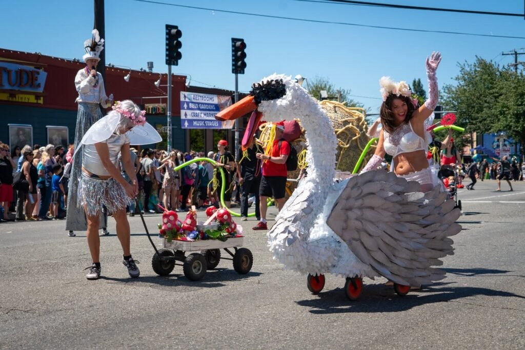 A large goose float rolls down a sunny parade street while whimsical performers wave to spectators.
