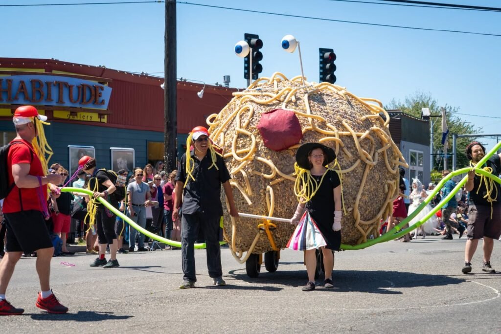 A large parade float shaped like a yellow noodle figure with googly eyes and green noodle legs. Spectators line the street during a sunny outdoor festival.
