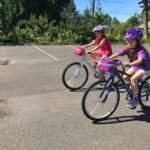 Two friends wearing helmets ride bicycles around an empty lot surrounded by green trees on a sunny day in the Pacific Northwest.