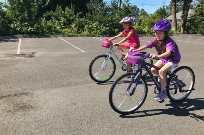 Two friends wearing helmets ride bicycles around an empty lot surrounded by green trees on a sunny day in the Pacific Northwest.