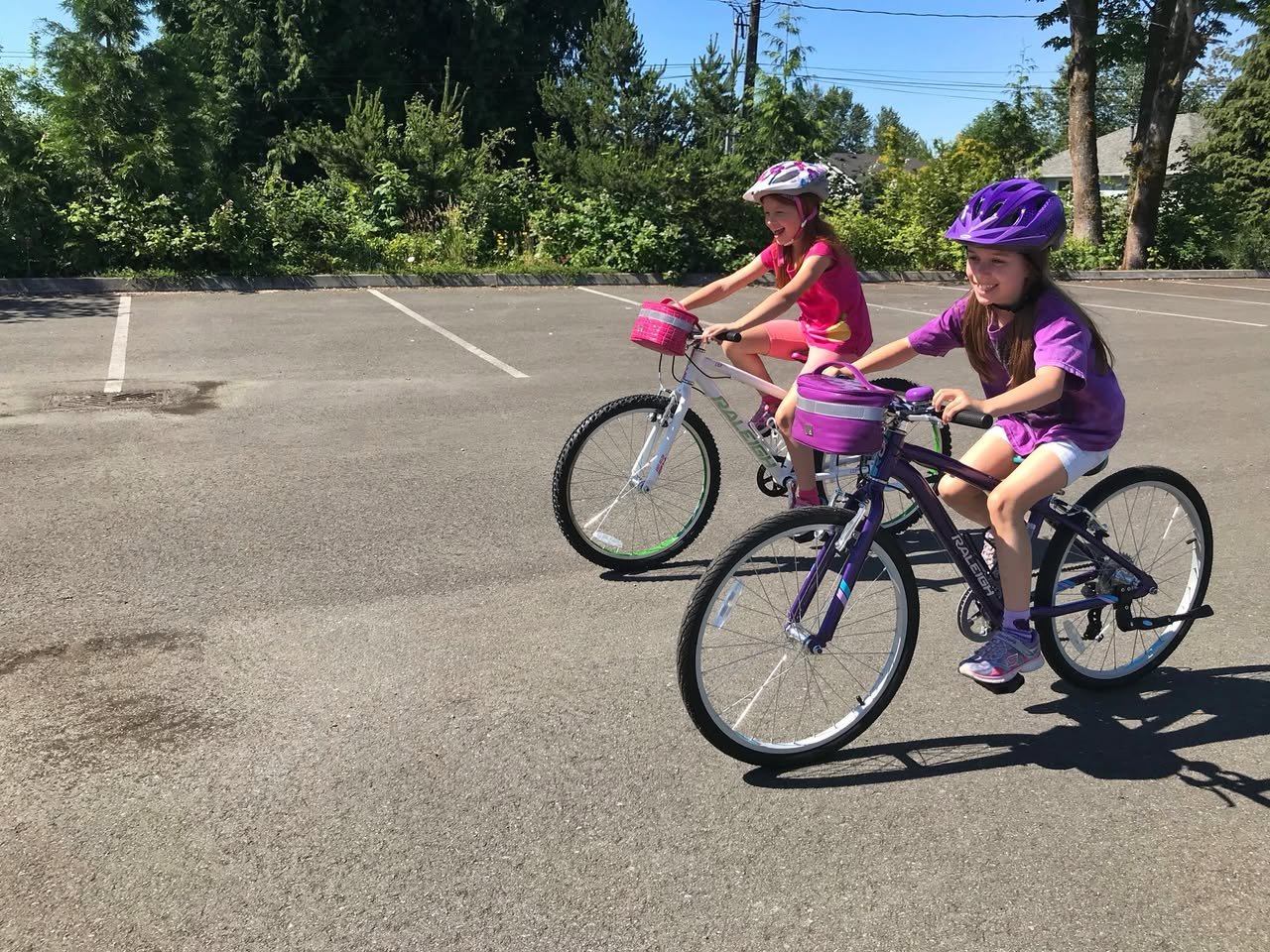 New Bikes Two friends wearing helmets ride bicycles around an empty lot surrounded by green trees on a sunny day in the Pacific Northwest.